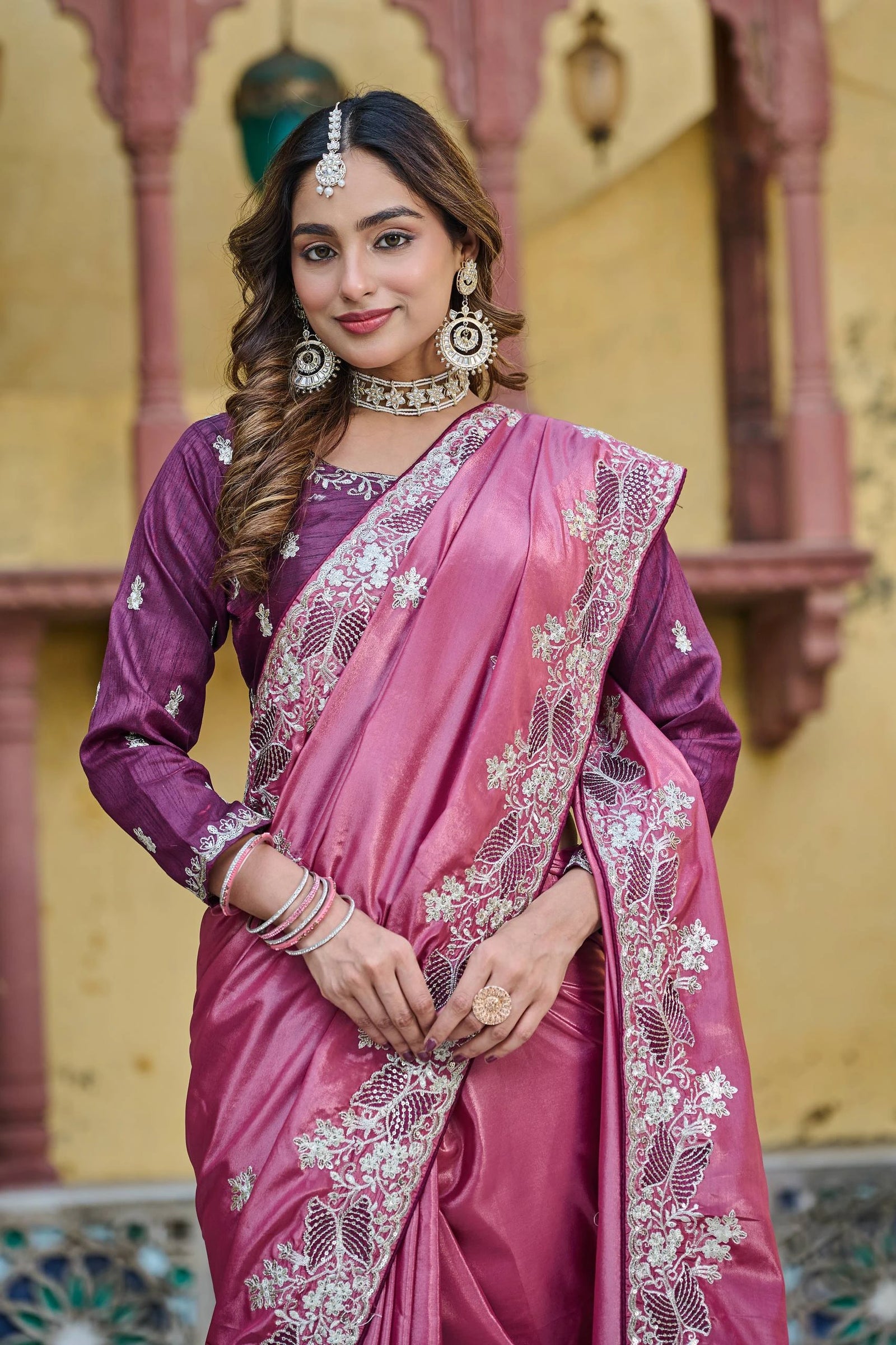 Woman in a pink and purple saree with intricate patterns, standing against a decorative architectural background.