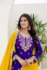 Woman wearing a purple and yellow traditional outfit with floral patterns, standing against a white wall with green plants.