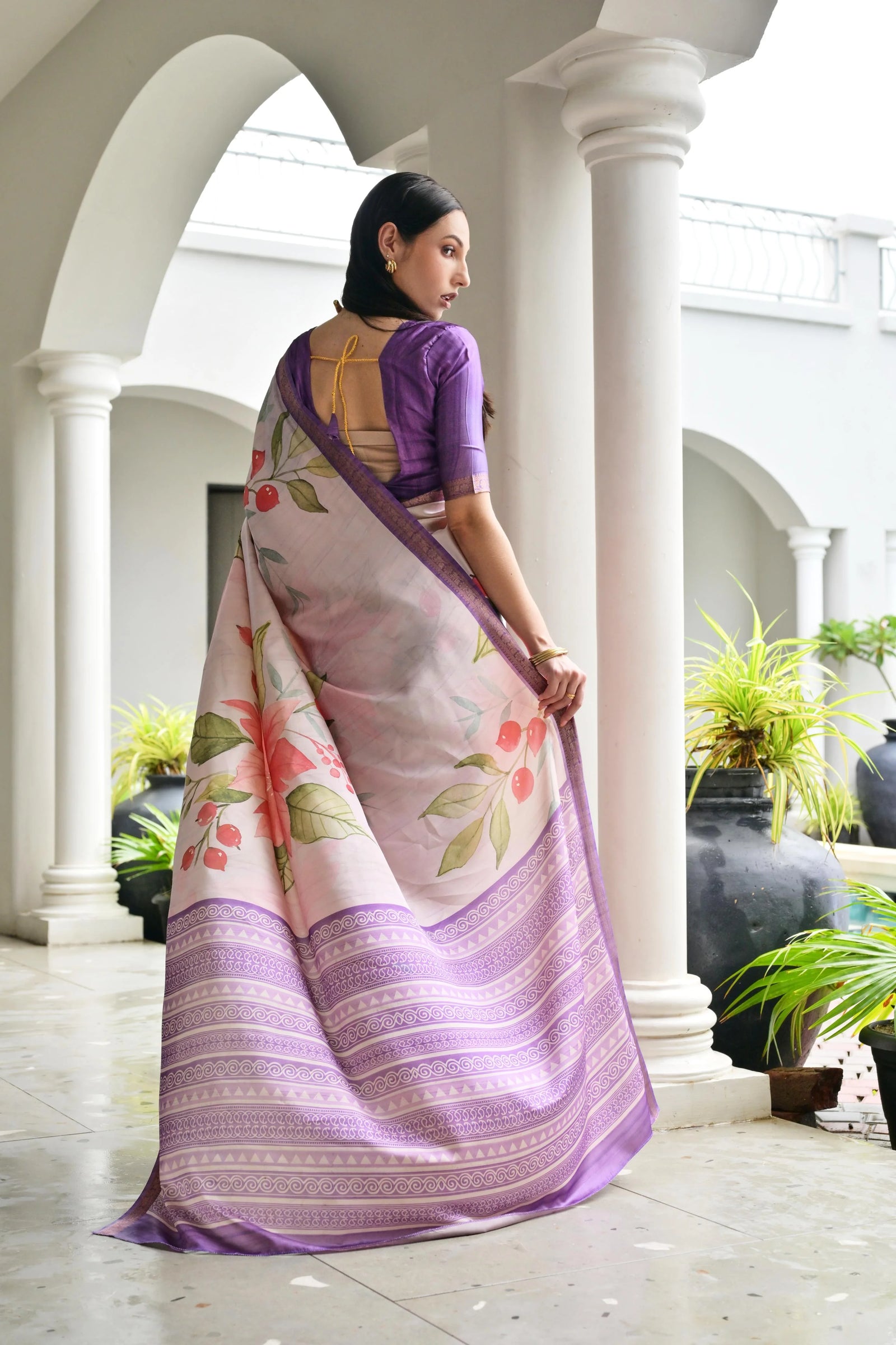 Woman in a floral saree standing in a decorative indoor setting with columns and plants.