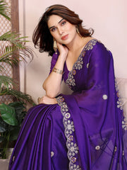 Woman wearing a purple saree with intricate designs, sitting indoors.