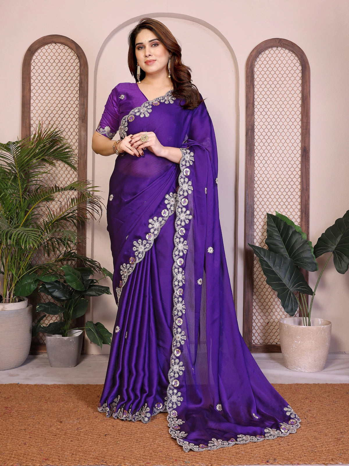 Woman wearing a purple saree with floral patterns in an indoor setting with plants.