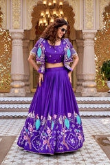 A full-length portrait of the woman in the indigo purple lehenga standing centered in a sunlit courtyard. The voluminous flare of the skirt is prominently displayed, showing the detailed repeating patterns of peacocks and flowering trees.
