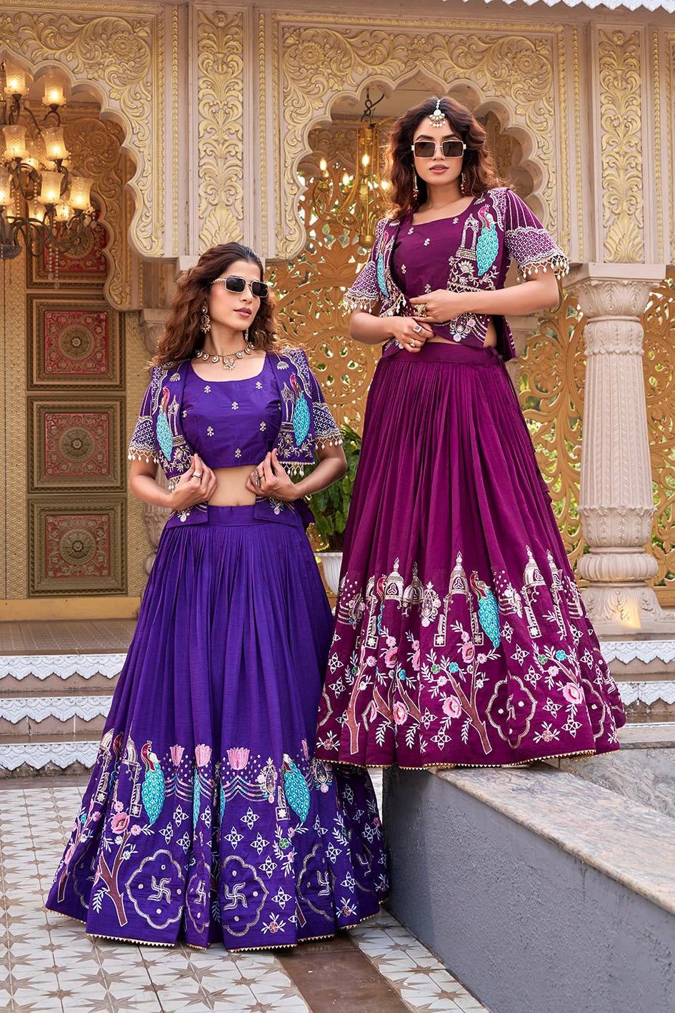 The women stand together under ornate golden arches. Both are wearing modern square sunglasses, blending contemporary styling with the traditional peacock-themed silk attire.