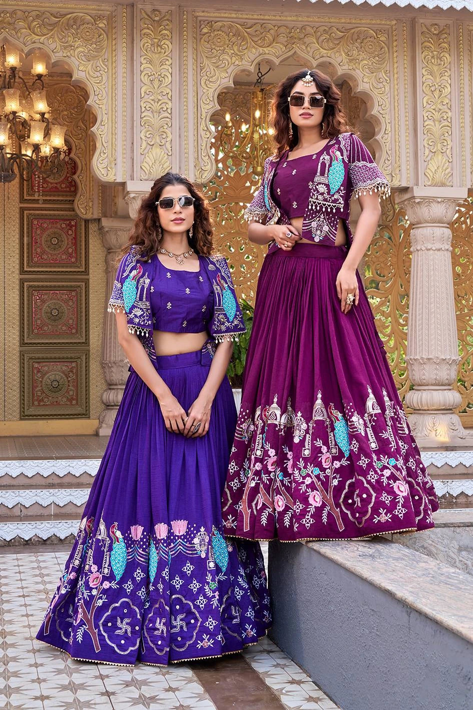 A full-length shot of two women posing in a traditional courtyard. One wears an indigo purple lehenga and the other a deep magenta version. Both outfits feature pleated silk skirts with elaborate peacock and floral embroidery, paired with a matching crop top and a short jacket with pearl-drop fringe