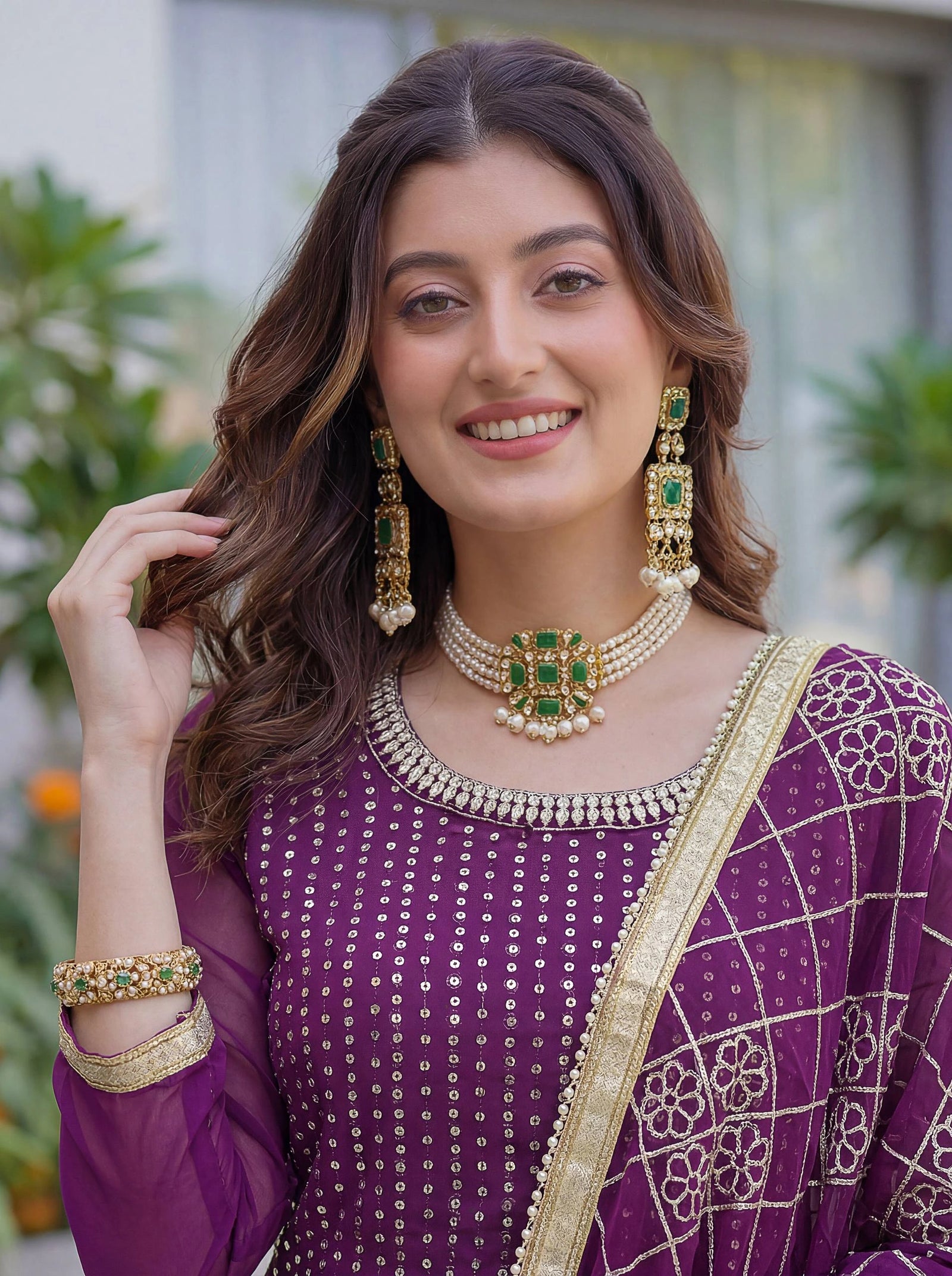 Woman wearing a purple traditional outfit with green and gold jewelry, standing outdoors.