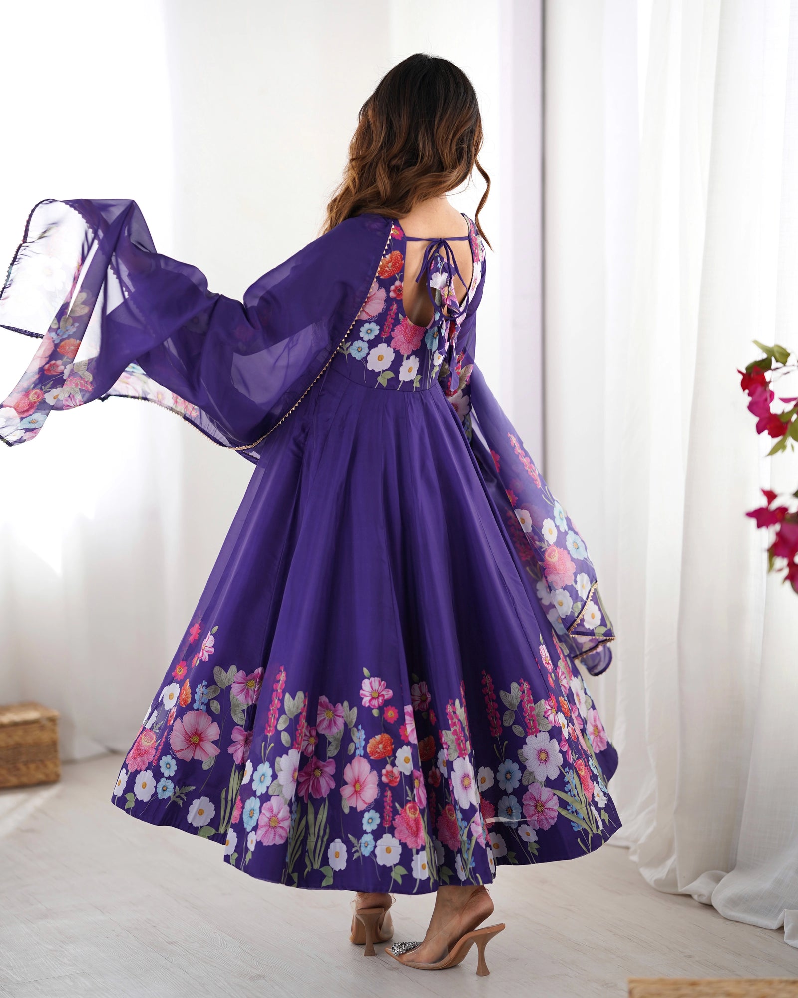 Woman wearing a purple floral dress with a sheer overlay against a white curtain background