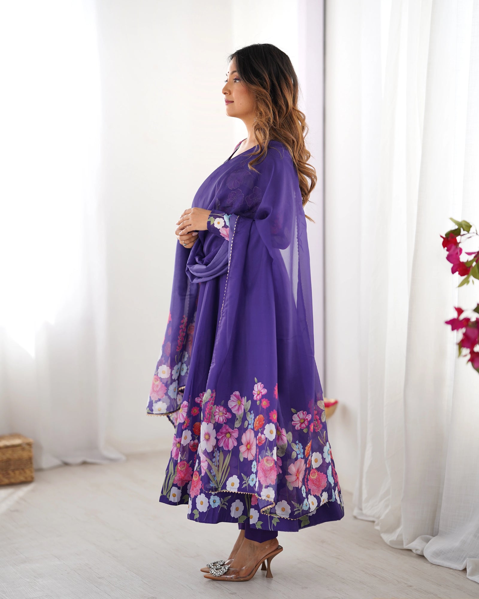 Woman wearing a purple floral dress standing in a bright room with white curtains.