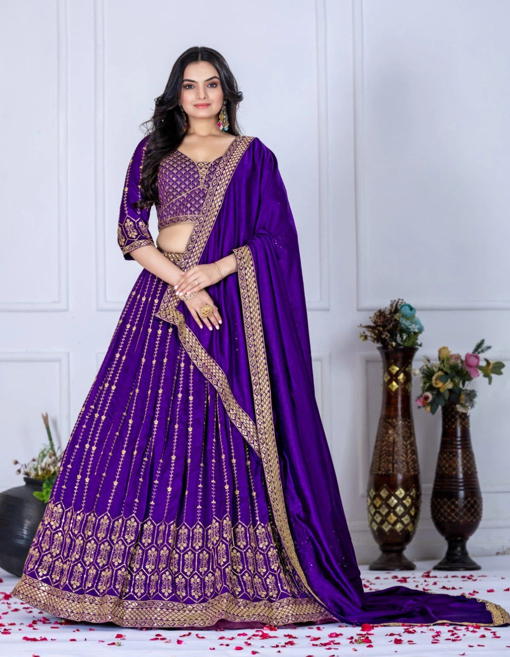 Woman wearing a purple saree with gold border against a white wall.