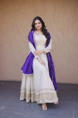 Woman in a white and purple traditional outfit standing against a beige wall.