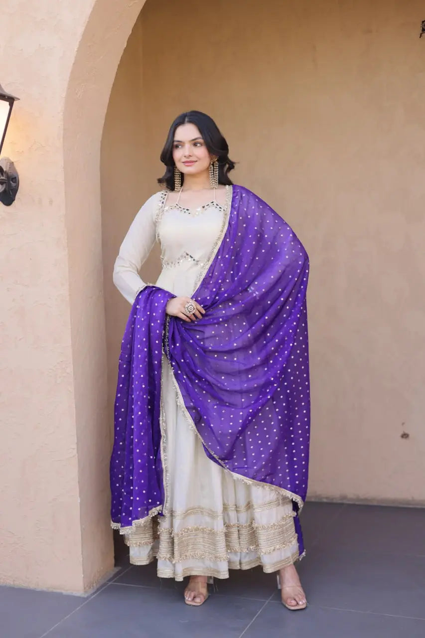 Woman wearing a white traditional outfit with a purple dupatta in front of a beige wall.