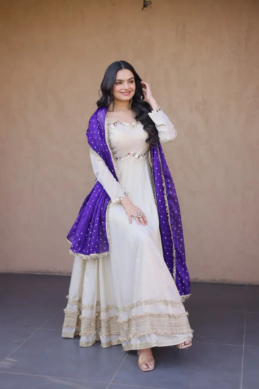 Woman in a white traditional outfit with a purple dupatta against a beige wall.