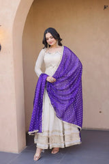 Woman wearing a white traditional outfit with a purple dupatta against a beige wall.