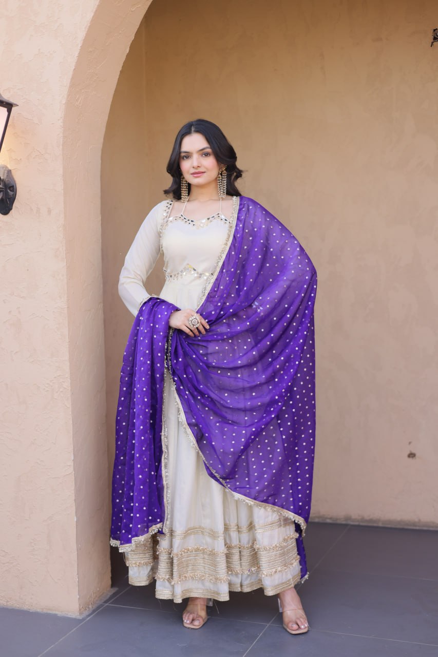 Woman wearing a white traditional outfit with a purple dupatta standing in front of a beige wall.