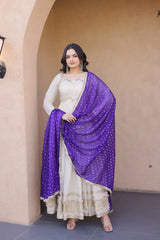 Woman wearing a white traditional outfit with a purple dupatta standing in front of a beige wall.