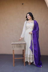 Woman in a white and purple traditional outfit standing next to a wooden stool against a beige wall.