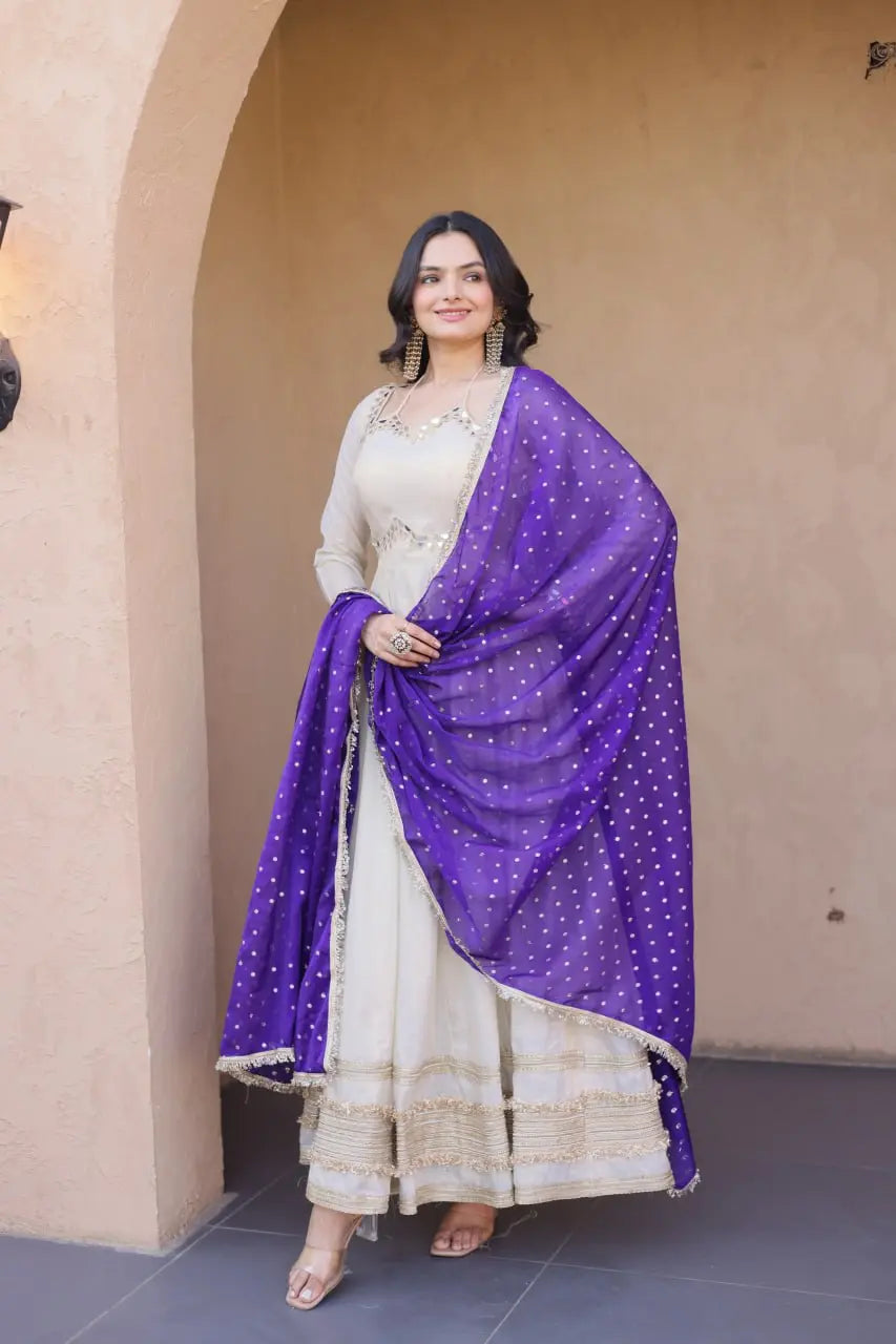 Woman in a white traditional outfit with a purple dupatta standing against a beige wall.