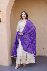 Woman in a white traditional outfit with a purple dupatta standing against a beige wall.