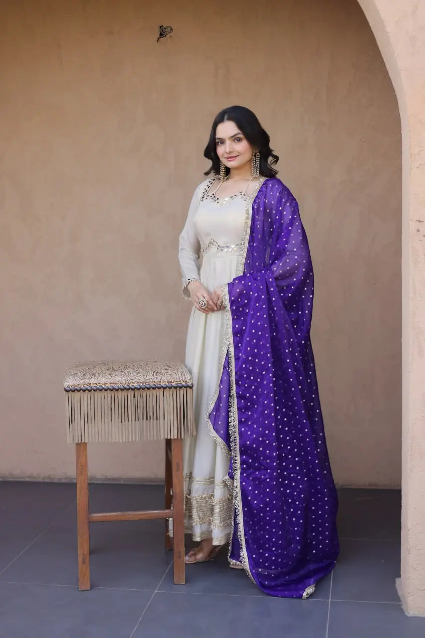 Woman in a white outfit with a purple dupatta standing next to a wooden stool against a beige wall.
