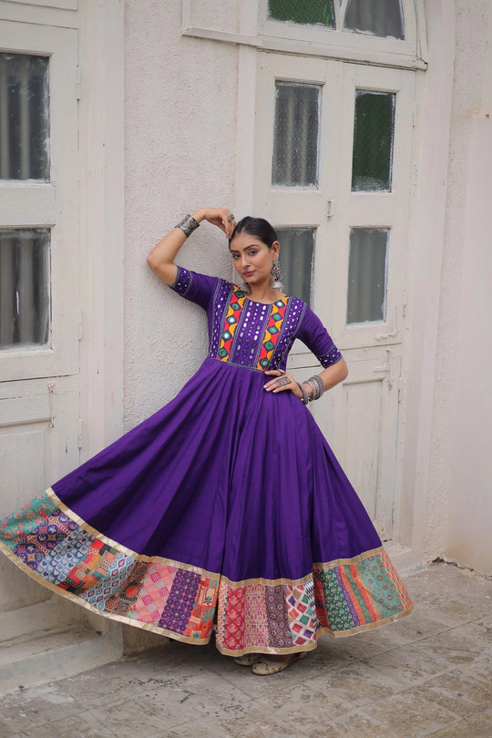 Woman in a vibrant purple traditional outfit with colorful patterns standing against a white wall.