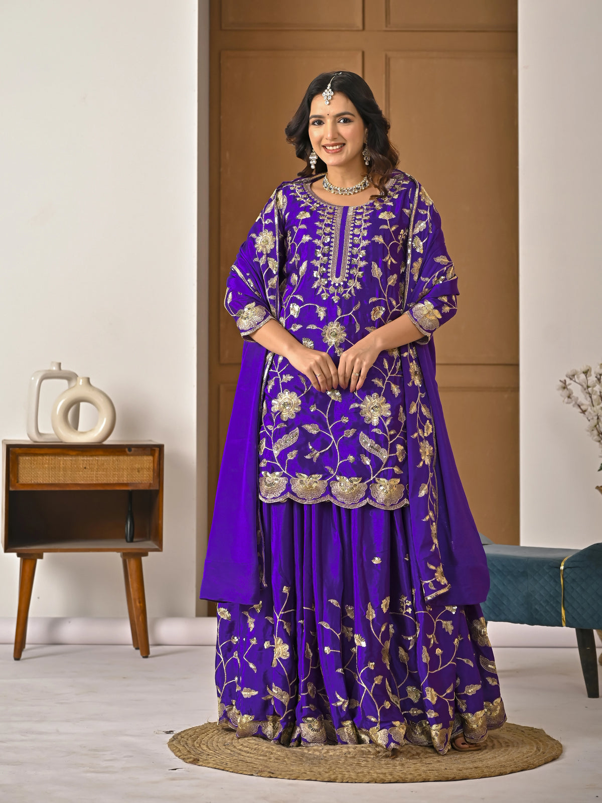 Woman in a purple embroidered traditional outfit standing in a room with wooden furniture and decor.