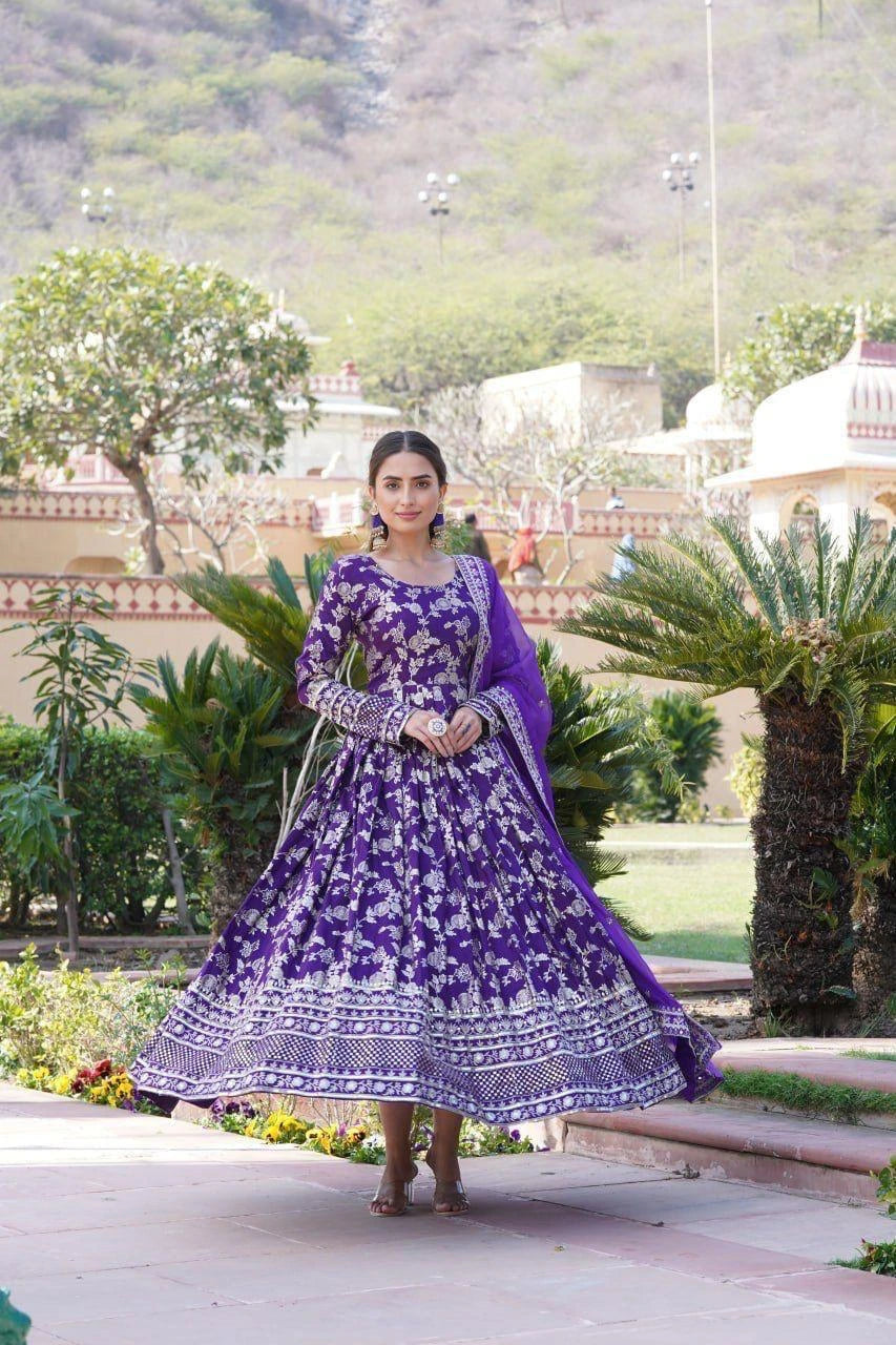 Woman in a purple floral dress standing outdoors with greenery and a building in the background