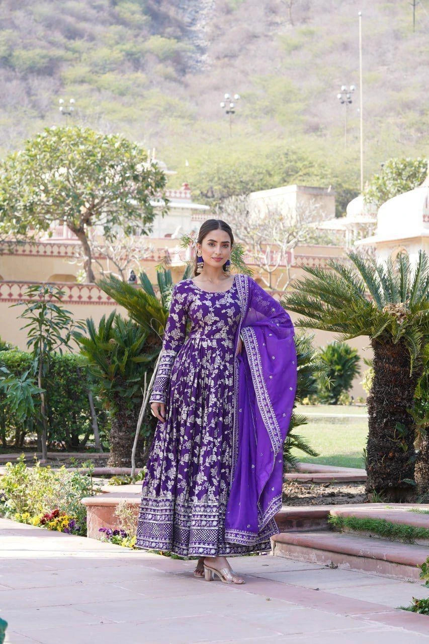 Woman in a purple traditional outfit standing in a garden with greenery and a building in the background