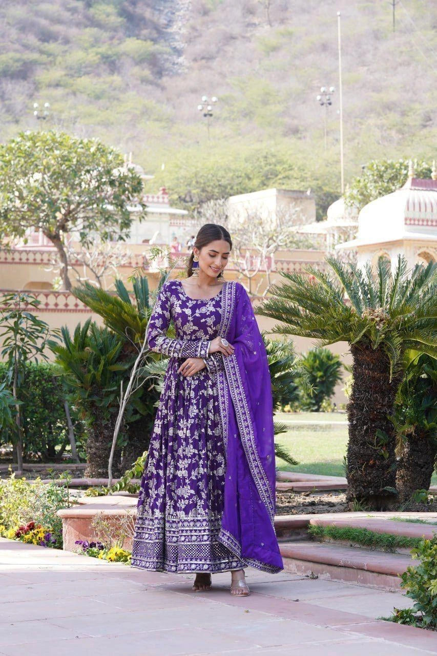 Woman in a purple traditional outfit standing in a garden with greenery and architectural elements in the background.