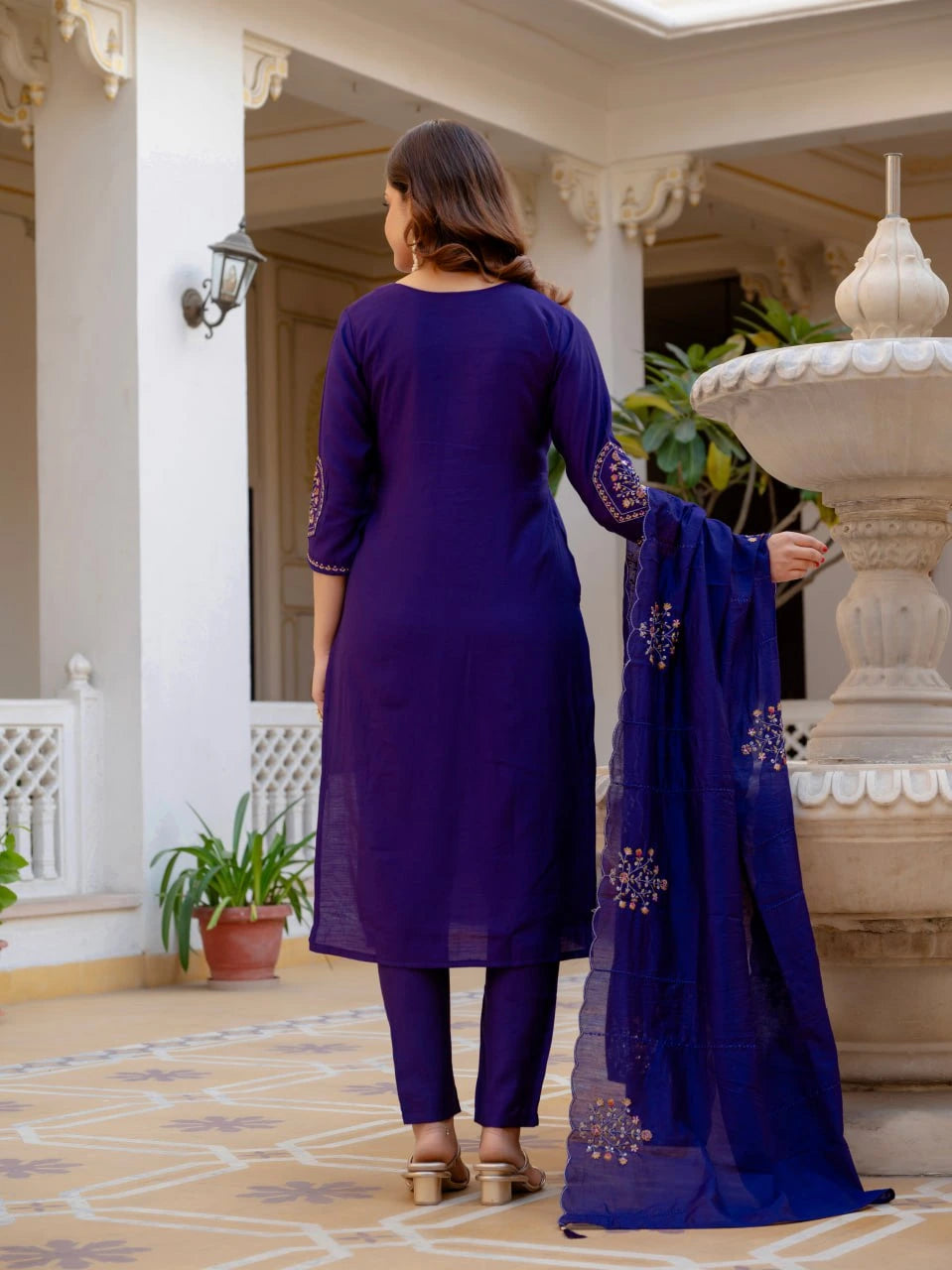 Woman in a purple traditional outfit standing in an elegant indoor setting with a fountain.