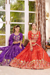Two women in colorful traditional outfits sitting indoors with a window and decorative curtains in the background.