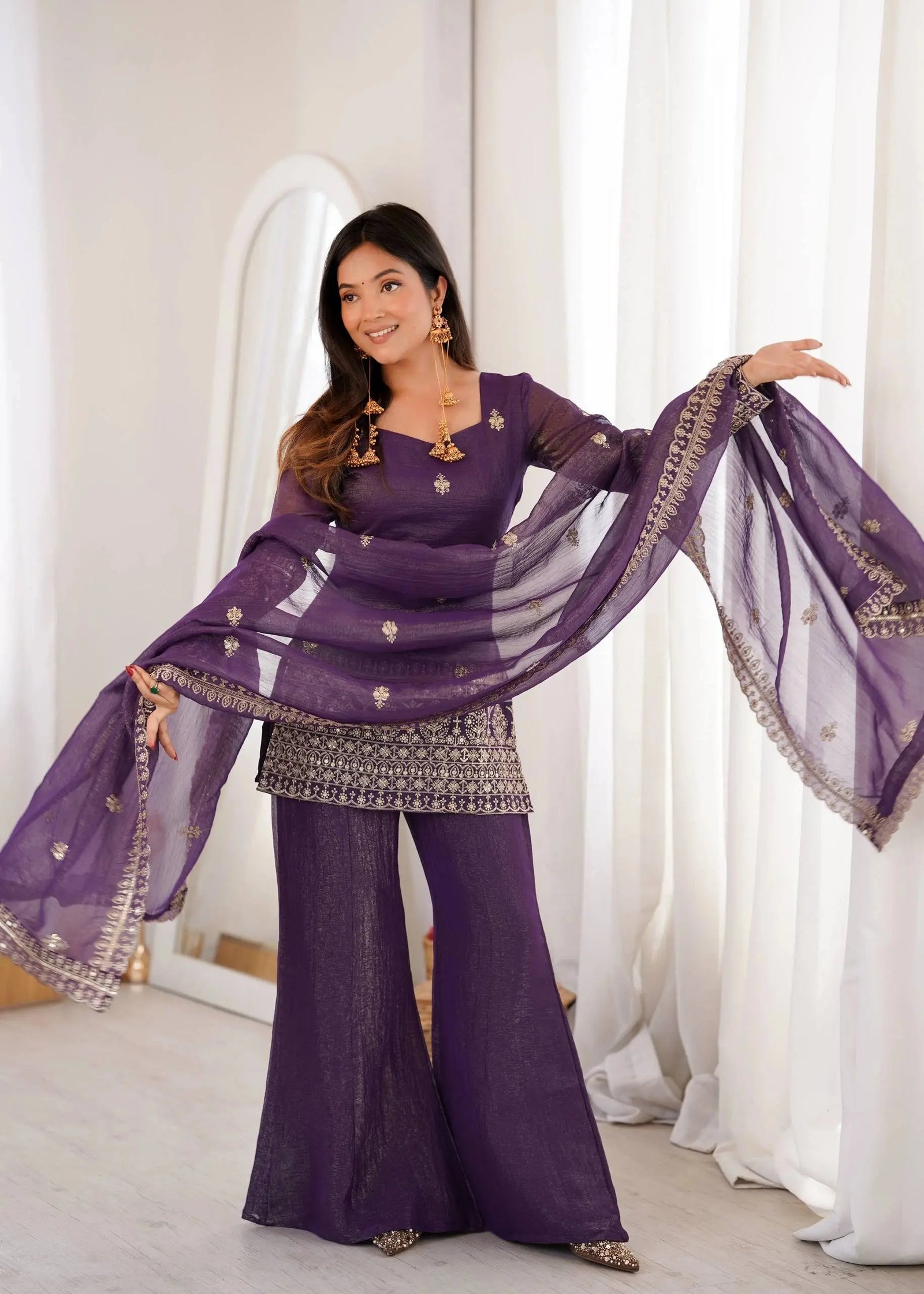 Woman in a purple traditional outfit with a sheer dupatta against a white curtain background