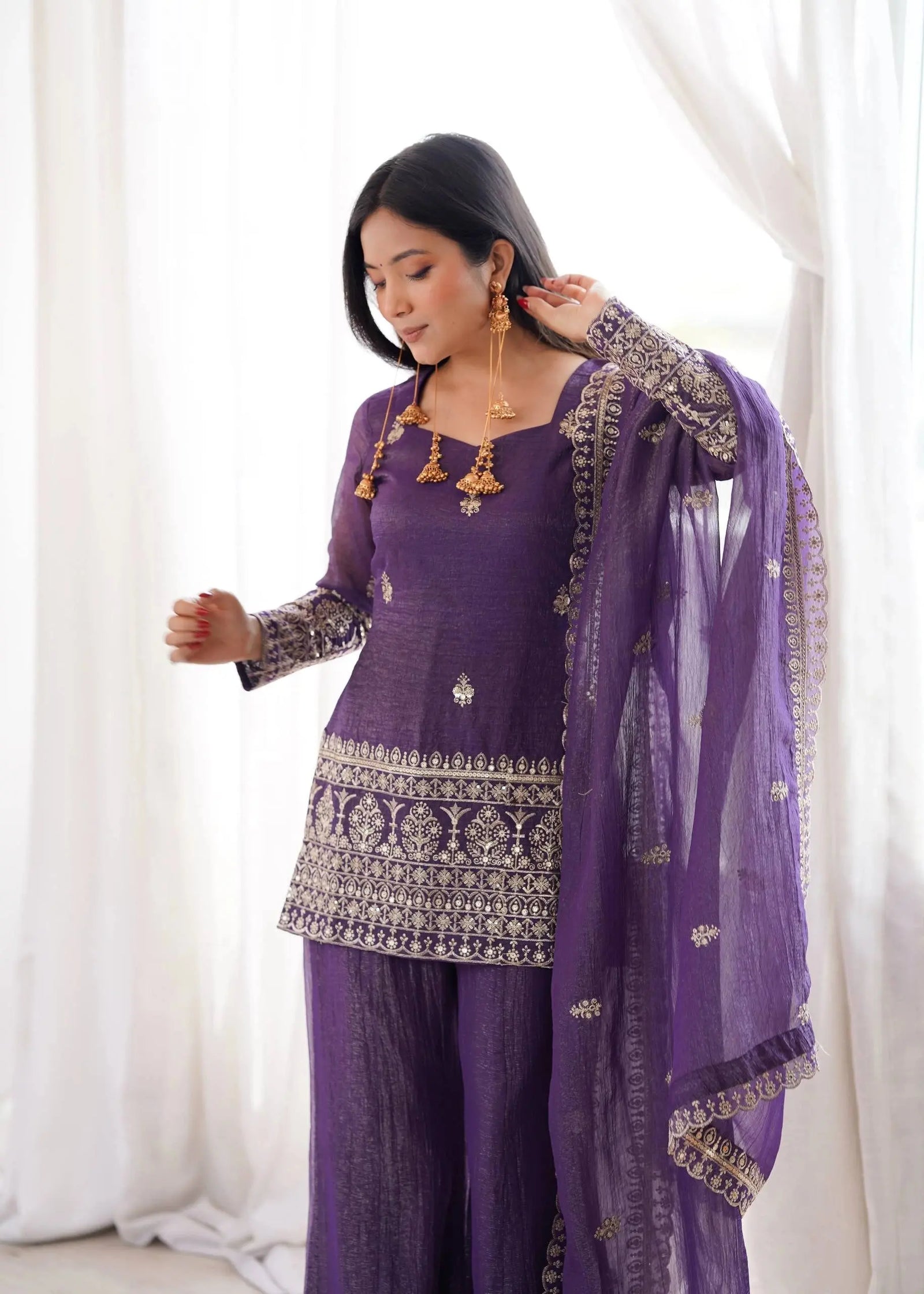Woman wearing a traditional purple outfit with intricate designs against a white curtain background