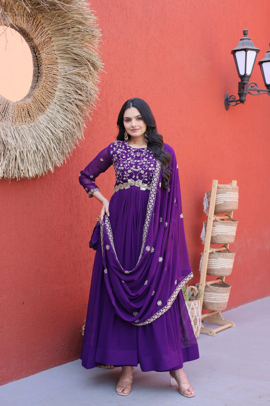 Woman in a purple traditional outfit standing against a red wall with decorative elements.