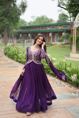 Woman in a purple dress with floral embellishments standing on a wooden path with greenery in the background.