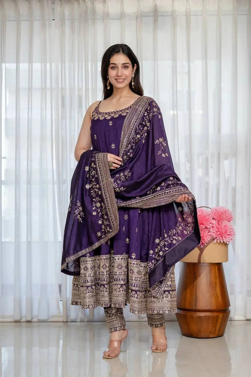 Woman in a traditional purple and gold outfit standing in front of a white curtain with pink flowers on a wooden stool.