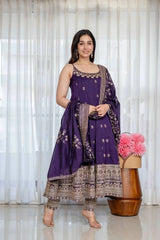 Woman in a purple and brown traditional outfit standing next to a wooden stool with pink flowers against a white curtain background.