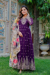 Woman in a purple dress with floral shawl standing outdoors against a decorative wall.