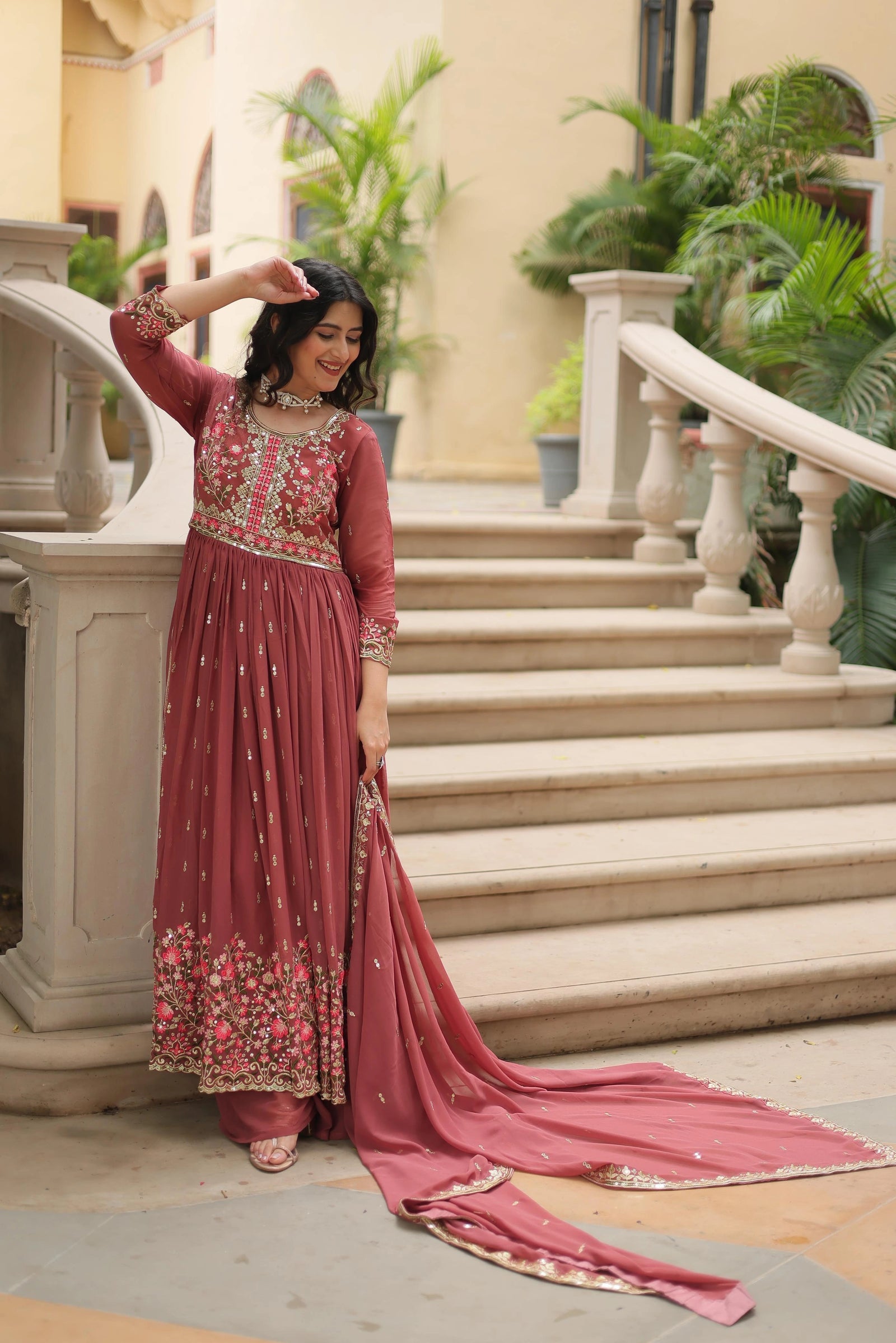 Woman in a traditional pink embroidered outfit standing on steps with decorative elements.