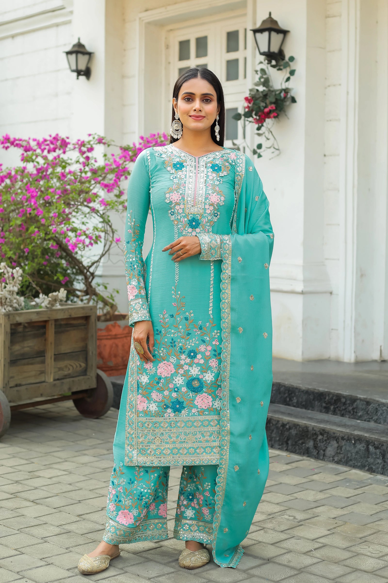 Woman in a turquoise embroidered traditional outfit standing in front of a white building with flowers.