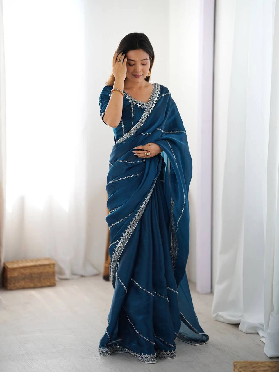 Woman wearing a blue saree with white patterns in a room with white curtains.