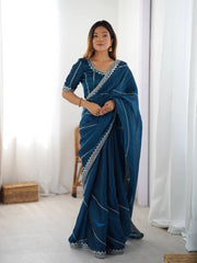 Woman wearing a blue saree with white patterns in a room with white curtains.