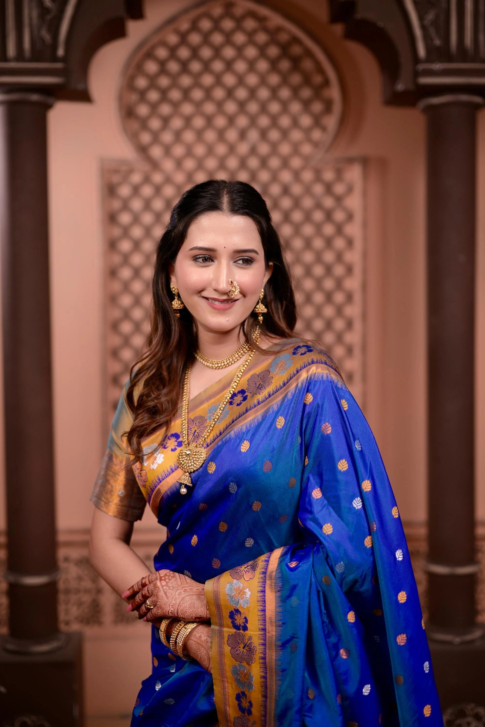 Woman in a blue saree with gold patterns standing in front of an ornate architectural background.