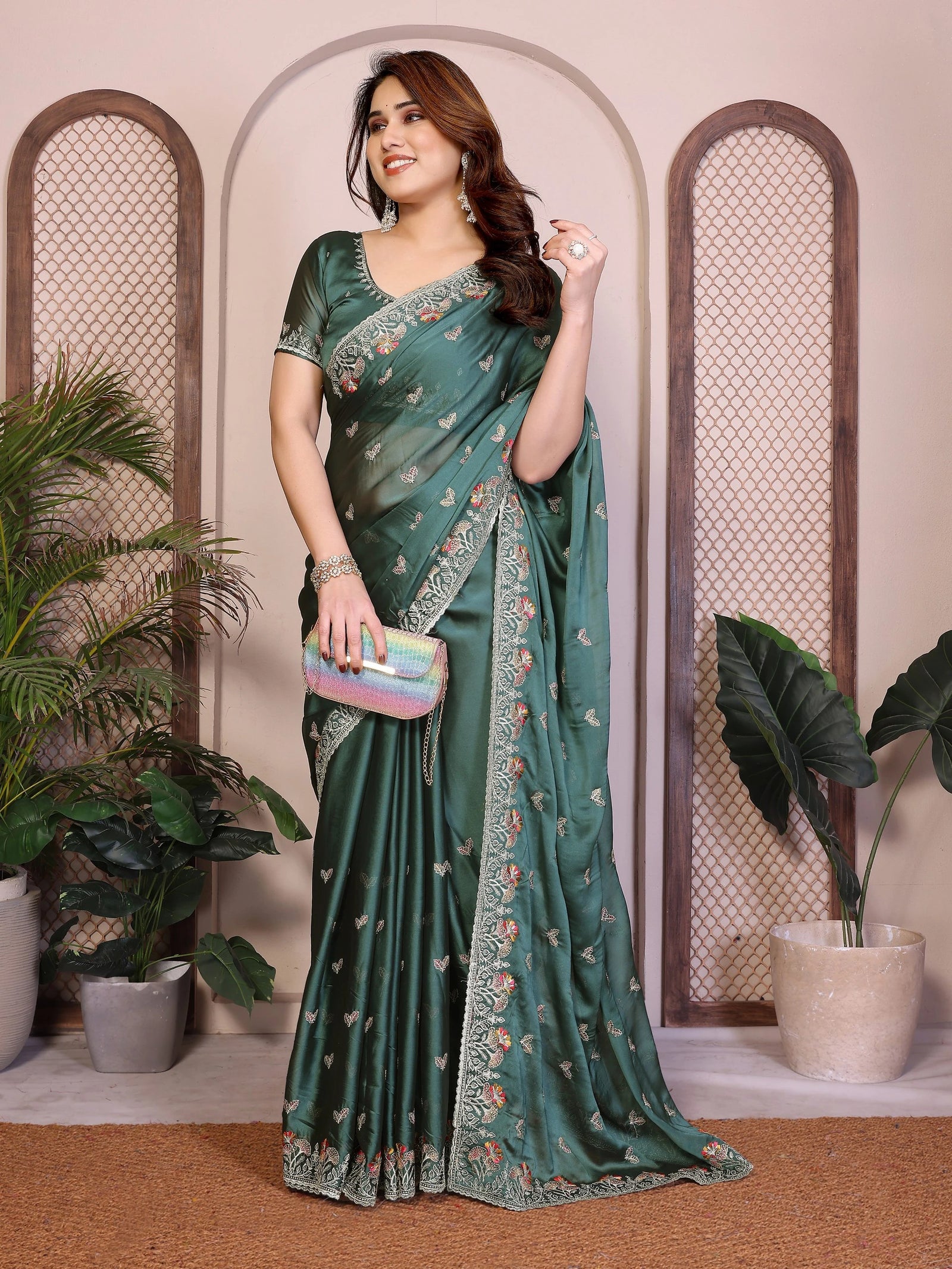 Woman in a green saree with floral patterns holding a colorful clutch, standing in front of decorative wall panels with plants.