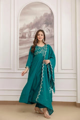 Woman wearing a teal embroidered dress standing in a room with white walls and a decorative mirror.