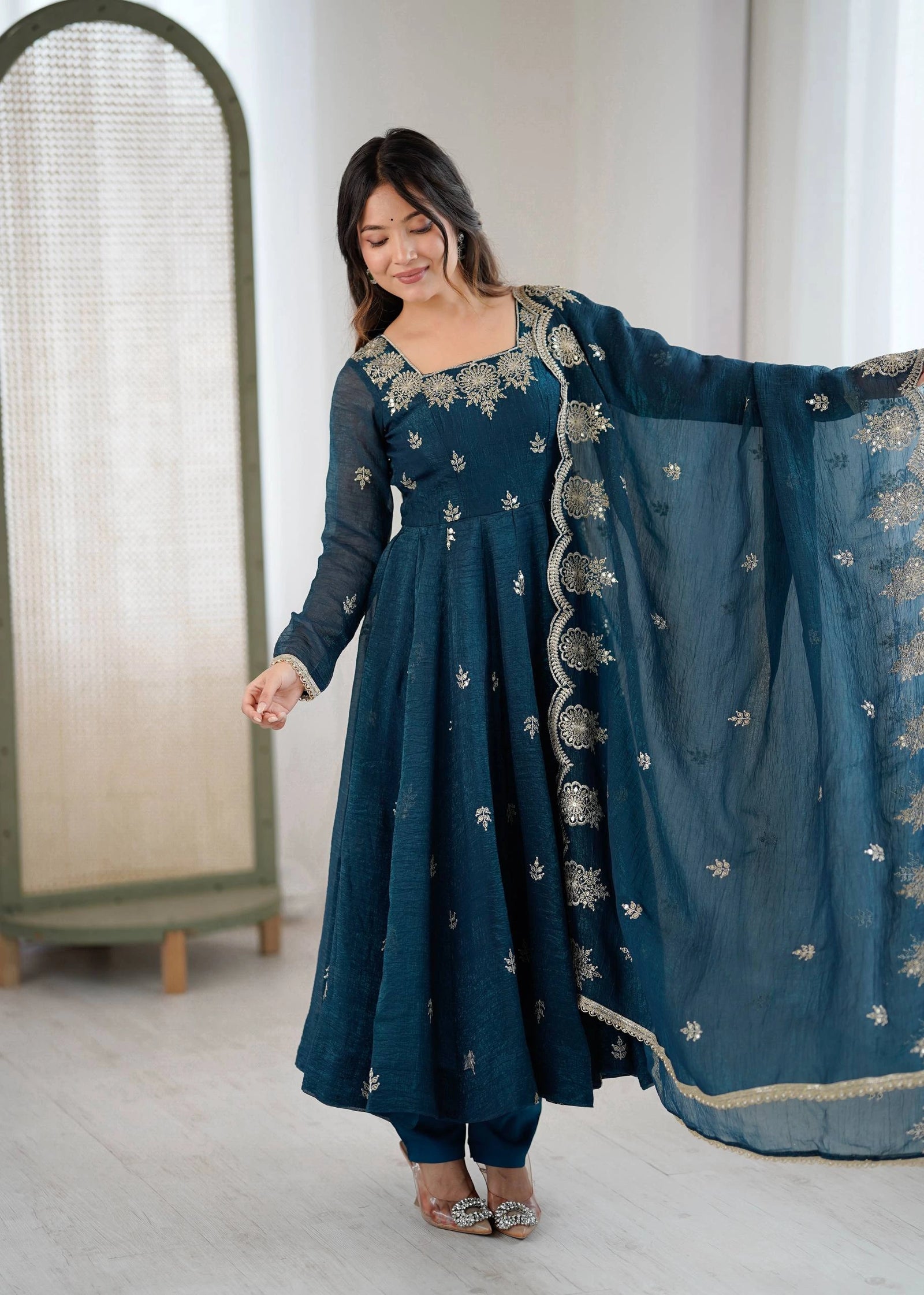 Woman wearing a teal embroidered outfit with a matching dupatta in a minimal indoor setting.