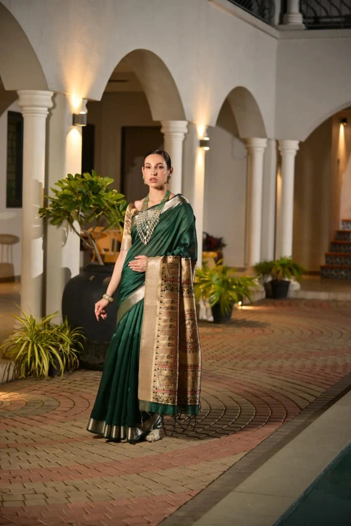 Woman in a green saree with gold border standing in an elegant indoor setting.