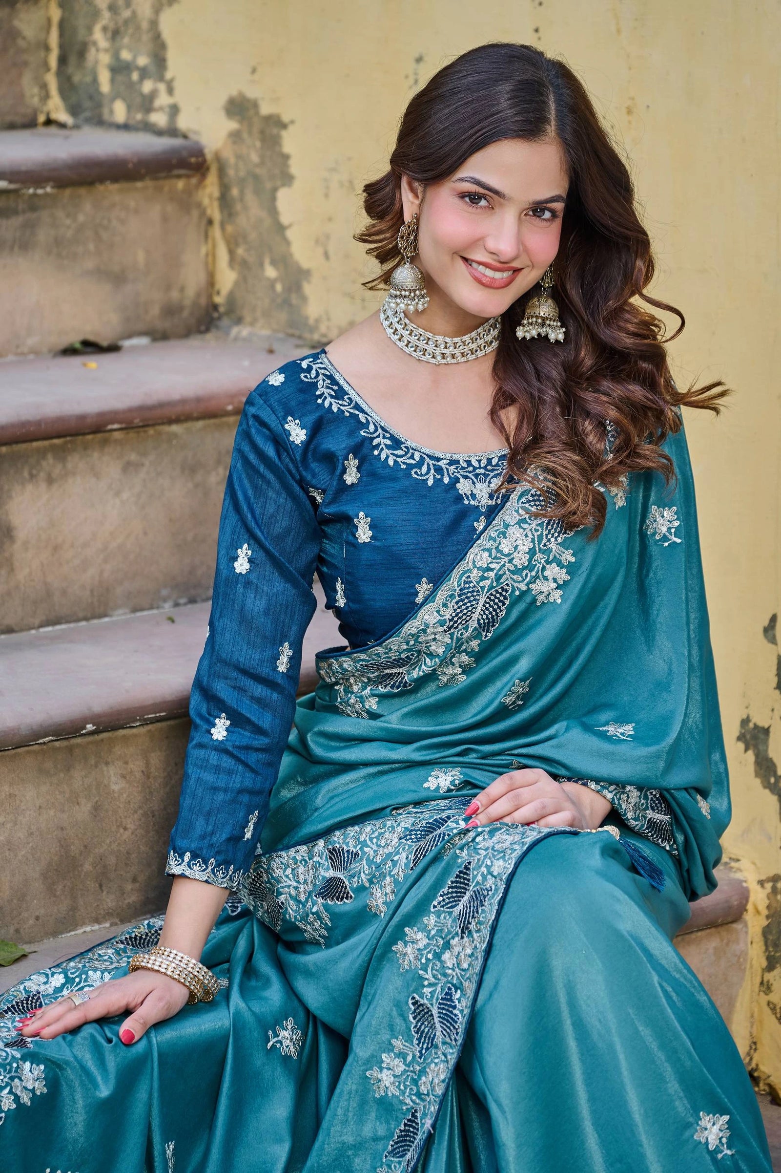 Woman in a blue saree sitting on steps with a yellow wall background