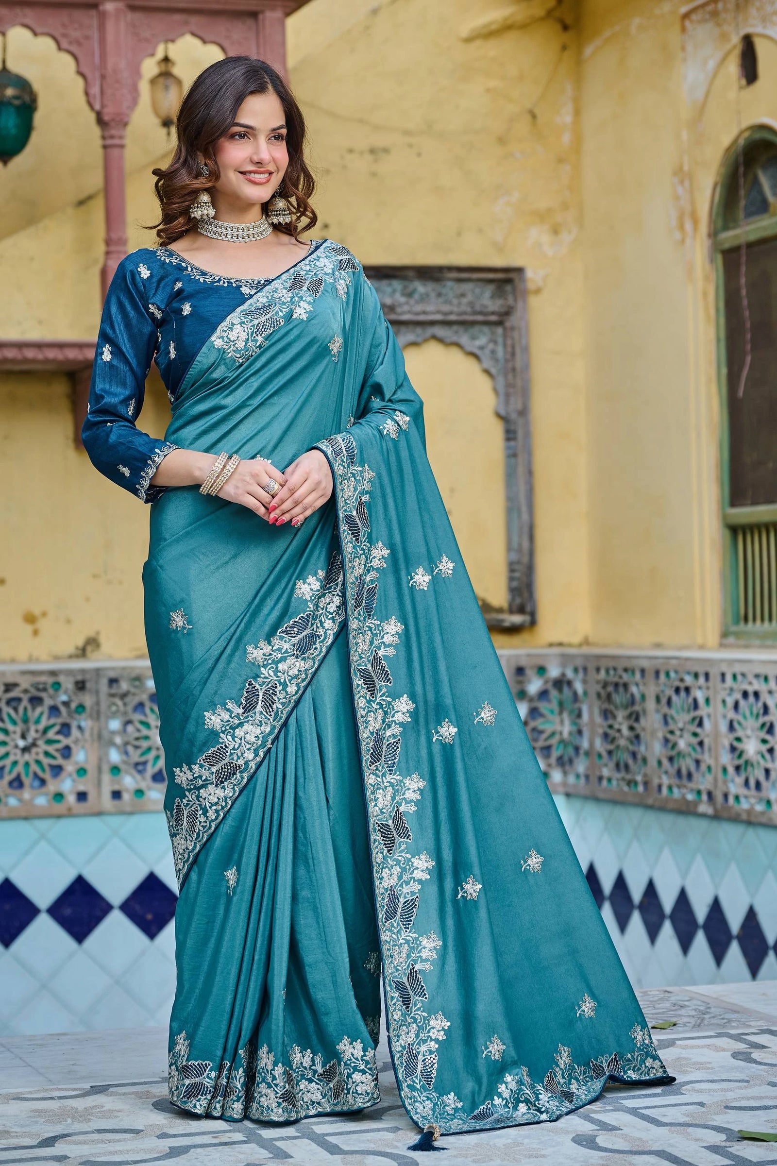 Woman wearing a teal saree with silver embroidery in front of a decorative building.