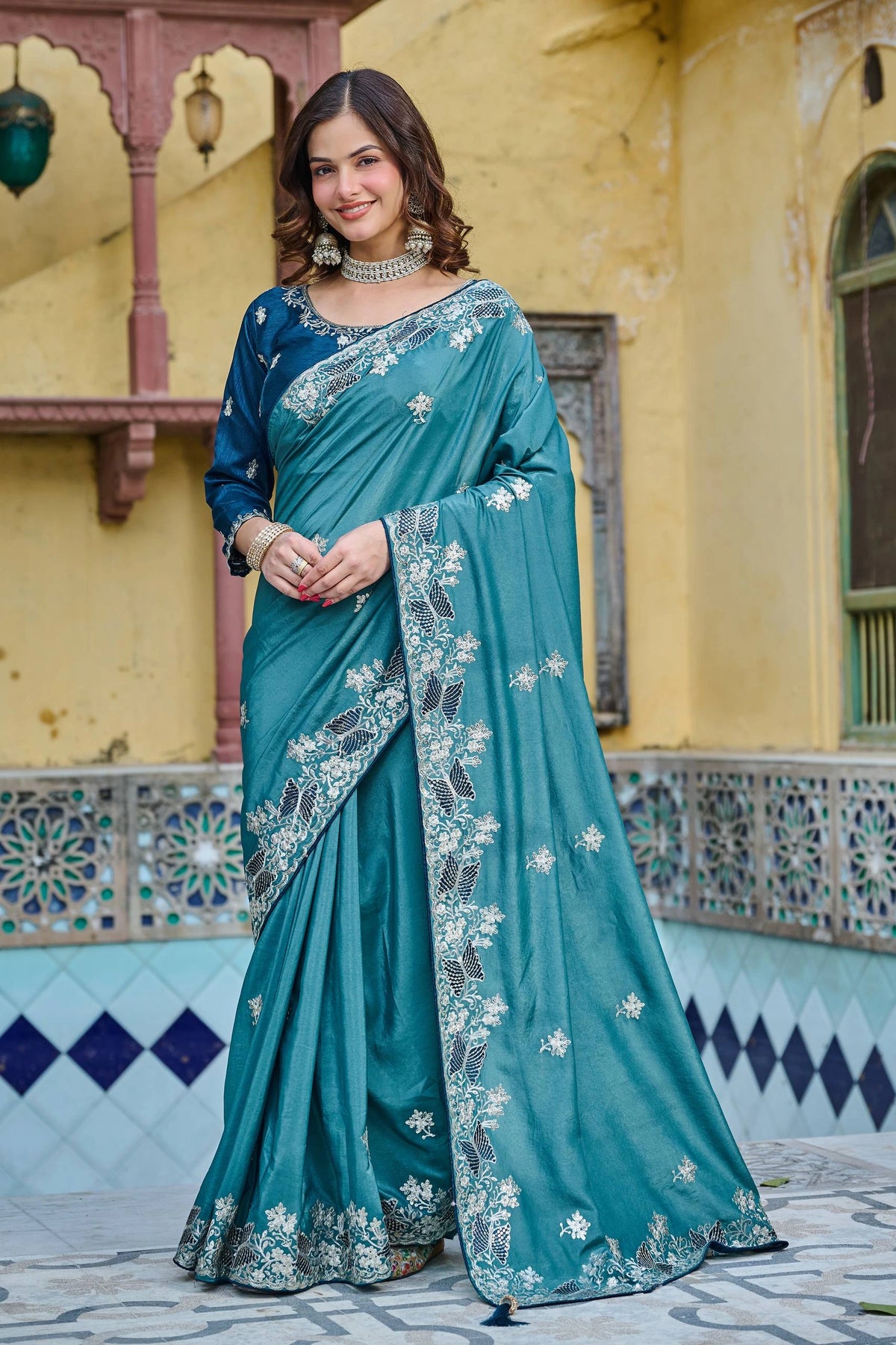 Woman wearing a teal saree with white floral patterns in front of a yellow building.