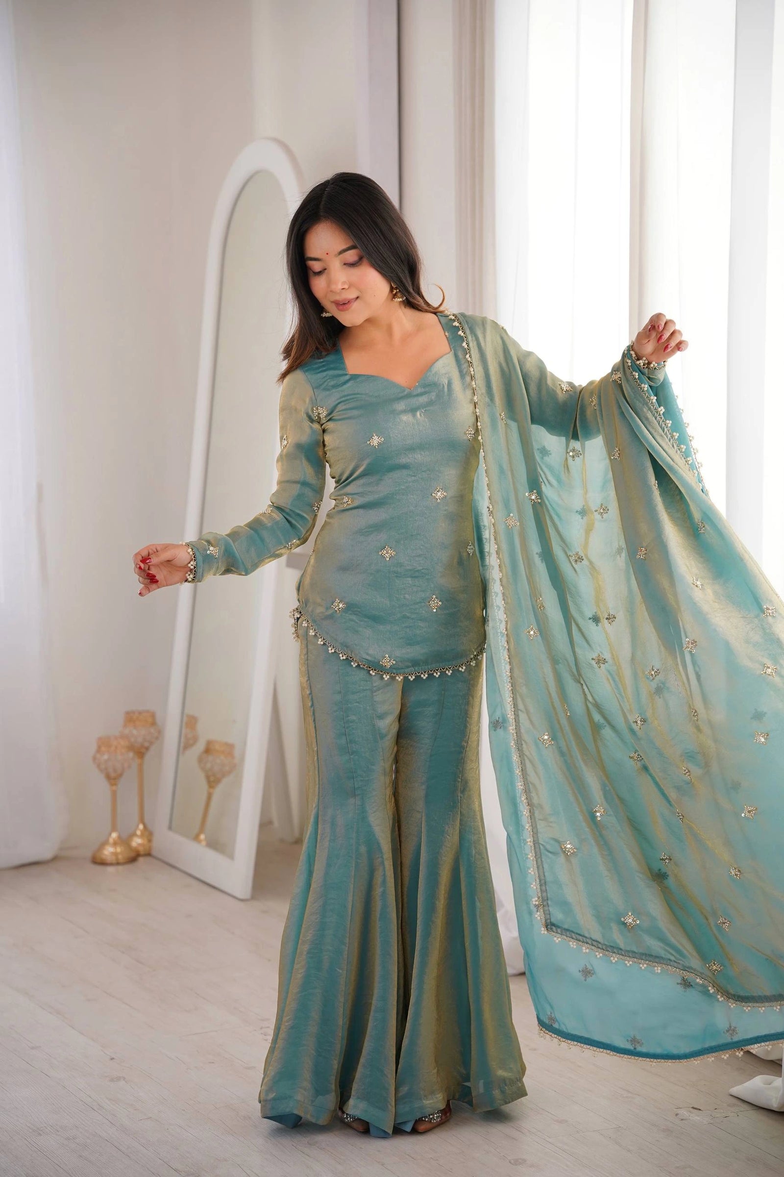 Woman in a teal traditional outfit standing in a bright room with white walls and a mirror.