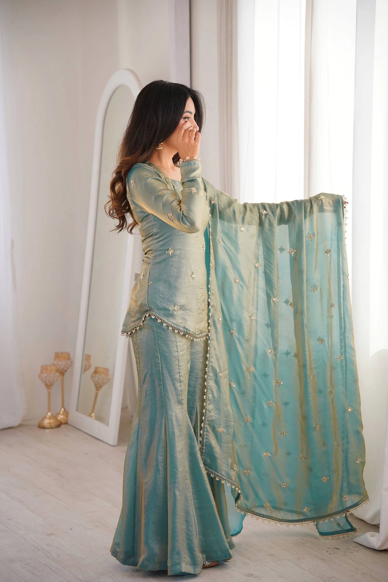 Woman in a light blue traditional outfit standing in a room with a mirror and window.